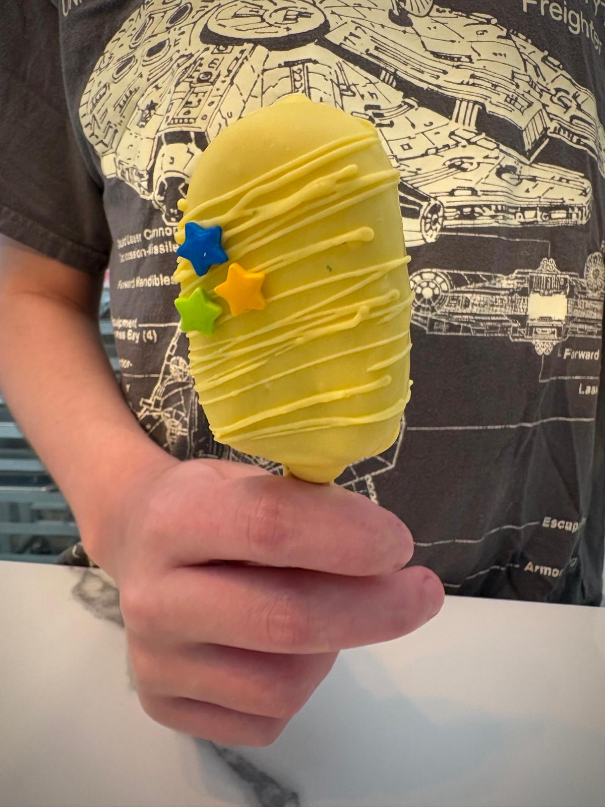 Hand holding a bright yellow cakesicle with aqua decoration